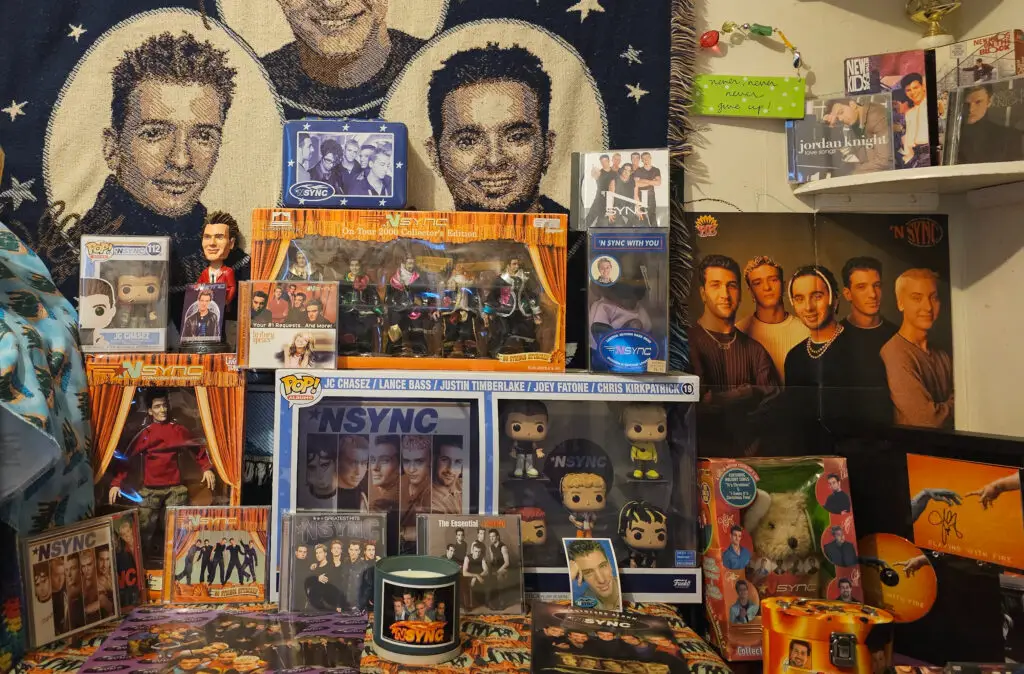 A colorful display of *NSYNC memorabilia, featuring a throw blanket with the band members' faces, a signed JC Chasez CD, action figures, Funko Pop figures, posters, collectible CDs, and a themed candle arranged neatly on a shelf. This collection highlights a fan's love for the iconic boy band NSYNC and their music.